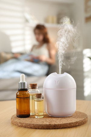 Aroma diffuser and bottles of essential oils on table, space for text. Woman reading book in room,...