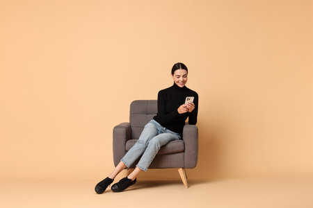 Smiling woman with smartphone sitting on armchair against beige backgroundの写真素材