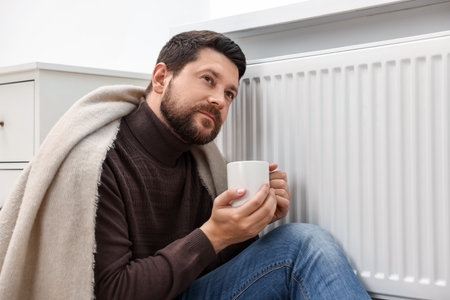 Man with cup of hot drink near heating radiator at home