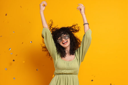 Happy young woman wearing carnival mask and confetti on yellow backgroundの写真素材