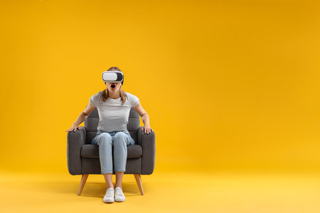 Emotional woman with virtual reality headset sitting in armchair on yellow background, space for textの写真素材
