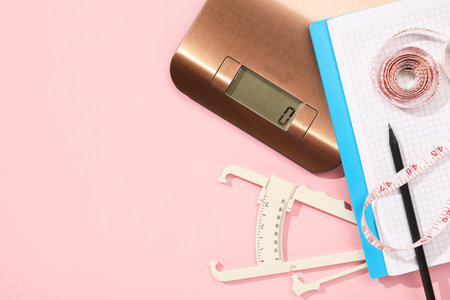 Body fat caliper, measuring tape, kitchen scale and notebook on pink background, flat lay. Space for textの写真素材