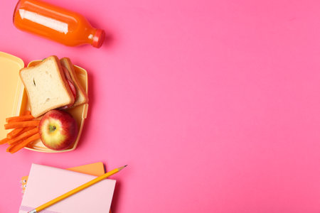 Lunch box with snacks, bottle of juice and stationery on pink background, flat lay. Space for textの写真素材