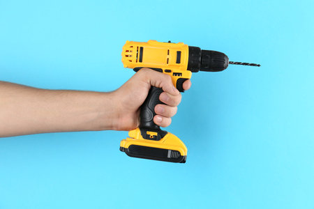 Man with cordless electric drill on light blue background, closeup