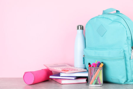 Backpack with different school stationery and bottle on light grey table against pink background....
