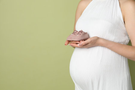 Young pregnant woman with pair of baby shoes on light olive background, closeup. Space for textの写真素材
