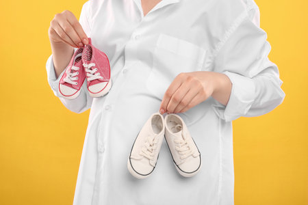 Expecting twins. Pregnant woman holding two pairs of shoes on yellow background, closeupの写真素材