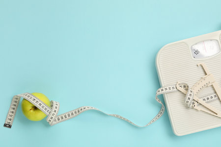 Body fat caliper, measuring tape, apple and notebook on light blue background, flat lay. Space for textの写真素材