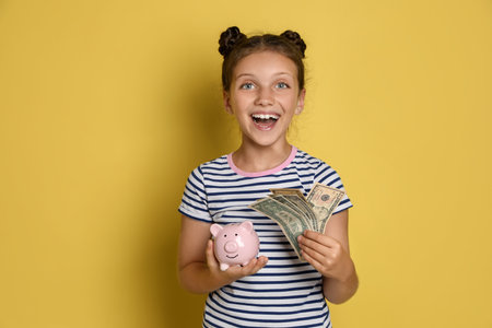 Pocket money. Cute girl with piggy bank and dollar banknotes on yellow backgroundの写真素材