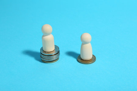 Financial inequality. One wooden figure on bigger stack of coins than another on light blue...