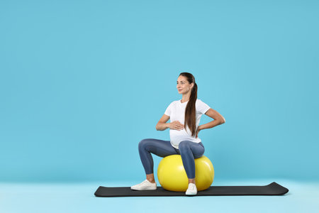 Beautiful pregnant woman doing exercises on fitball against light blue backgroundの写真素材