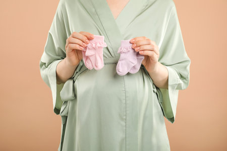 Expecting twins. Pregnant woman holding two pairs of socks on light brown background, closeupの写真素材