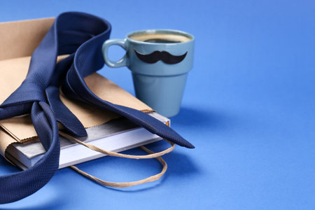 Happy Father's Day. Paper bag with book, tie and cup of coffee on blue background, closeup. Space...