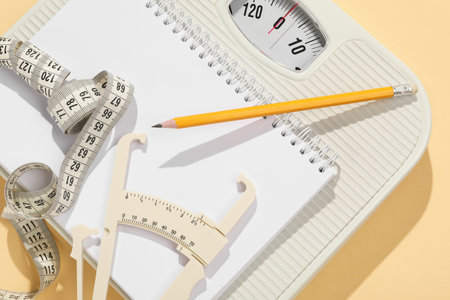 Body fat caliper, measuring tape, scale, pencil and notebook on beige background, top view