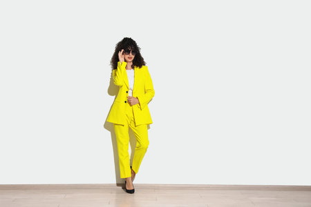 Beautiful young woman in stylish yellow suit and sunglasses near light wall, space for textの写真素材