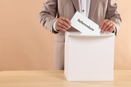 referendum. Man putting his vote into ballot box at wooden table against beige background, closeupの写真素材