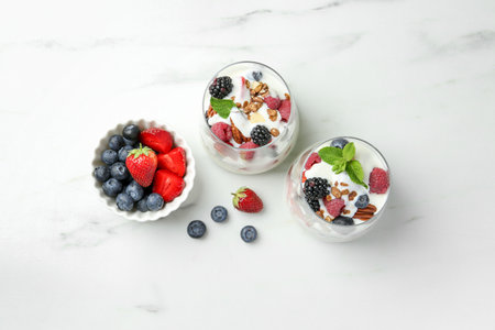 Tasty yogurt with fresh berries, granola and mint in glasses on white marble table, flat layの写真素材