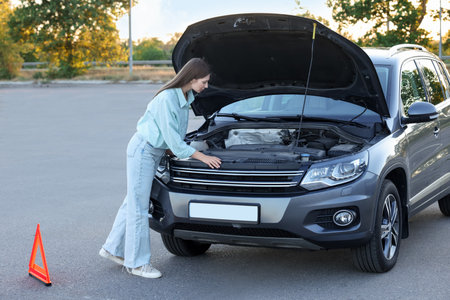 Stressed woman looking under hood of broken car outdoorsの写真素材