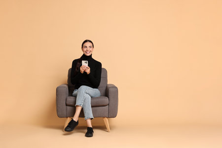 Smiling woman with smartphone sitting on armchair against beige background. Space for textの写真素材