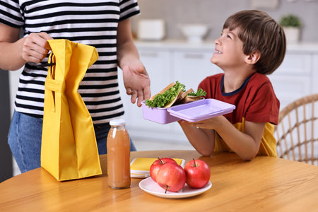 Mother and her cute son preparing school lunch box with healthy food at wooden table in kitchen, closeupの写真素材