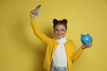 Pocket money. Cute girl with piggy bank and dollar banknotes on yellow backgroundの写真素材