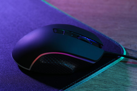 Black computer mouse and mousepad on wooden table in neon lights, closeupの写真素材