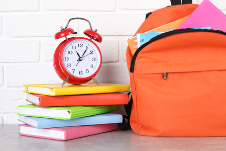 Backpack with different school stationery and alarm clock on light gray table near brick wall,...