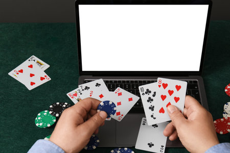 Online poker. Man holding playing cards and chip at green table with laptop, closeupの写真素材