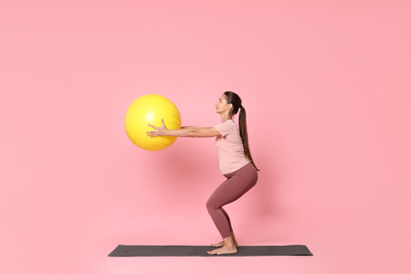 Beautiful pregnant woman with fitball doing exercises on mat against pink backgroundの写真素材
