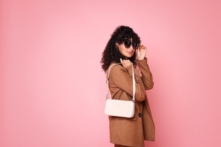 Beautiful young woman in stylish suit and sunglasses with bag on pink backgroundの写真素材