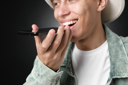 Young man recording voice message via smartphone on dark background, selective focusの写真素材