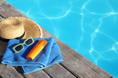 Stylish sunglasses, towel, hat and sunscreen near outdoor swimming pool. Space for text