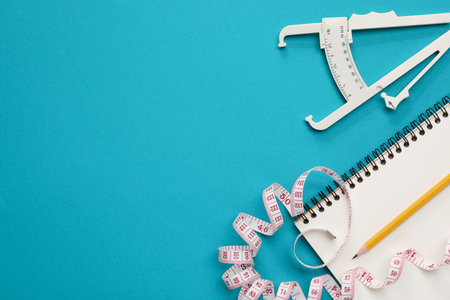 Body fat caliper, measuring tape, notebook and pencil on light blue background, flat lay. Space for textの写真素材