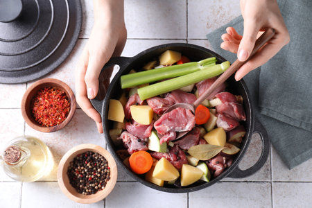 Woman preparing stew with vegetables and meat at white tiled table, top view