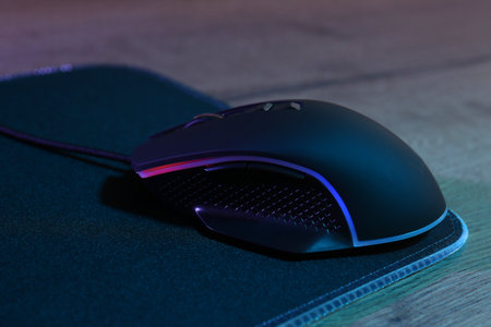 Black computer mouse and mousepad on wooden table in neon lights, closeupの写真素材