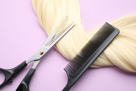 Blonde hair strand, comb and professional scissors on violet background, top view