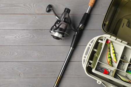Different fishing tackles in box and rod with spinning reel on gray wooden table, top view. Space...
