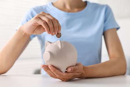 Woman putting coin into piggy bank at white table indoors, closeupの写真素材