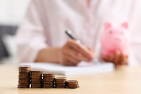 Woman with piggy bank taking notes at table indoors, focus on coinsの写真素材