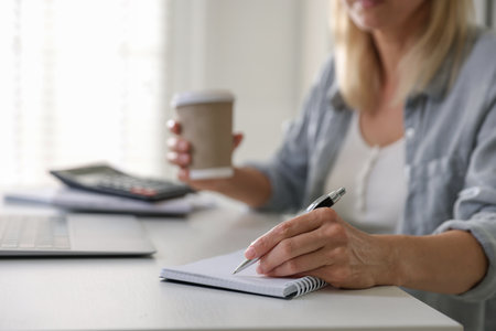 Budget planning. Woman with paper cup of taking coffee notes at table indoors, closeupの写真素材