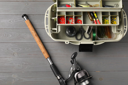 Different fishing tackles in box and rod with spinning reel on gray wooden table, top view. Space for textの写真素材