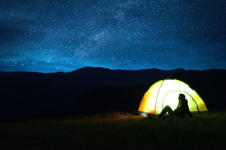 Woman sitting near modern camping tent in wilderness at night, space for textの写真素材