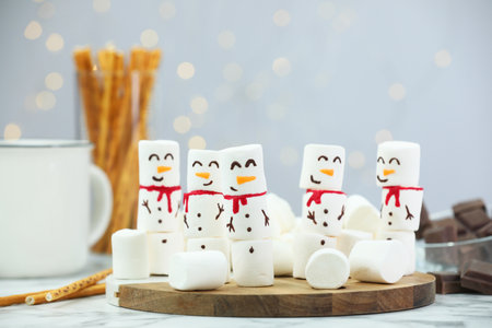 Funny snowmen made with marshmallows and other treats on white table against blurred lightsの写真素材
