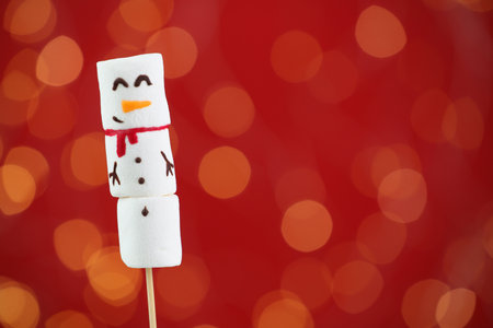 Funny snowman made of marshmallows on red background with blurred lights. Space for textの写真素材