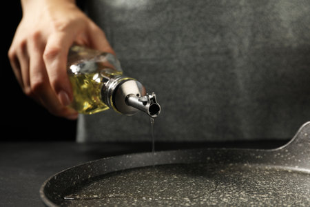 Woman pouring oil into frying pan at dark gray textured table, closeupの写真素材