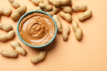 Tasty peanut butter in bowl and groundnuts on beige background, flat lay. Space for text