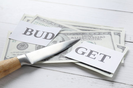 Cut paper with word Budget, dollar banknotes and knife on white wooden table, closeup