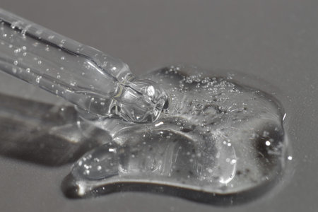 Dripping cosmetic serum from pipette on gray background, closeup