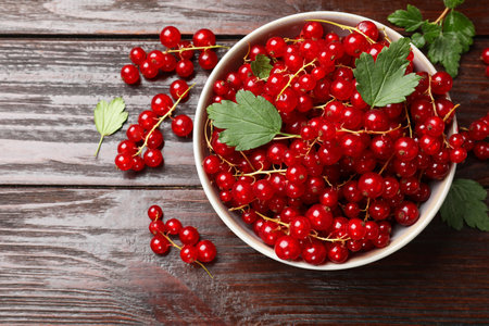 Fresh red currents in bowl and green leaves on wooden table, top view
