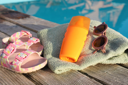 Stylish sunglasses, shoes, towel and sunscreen on wooden deck near outdoor swimming poolの写真素材